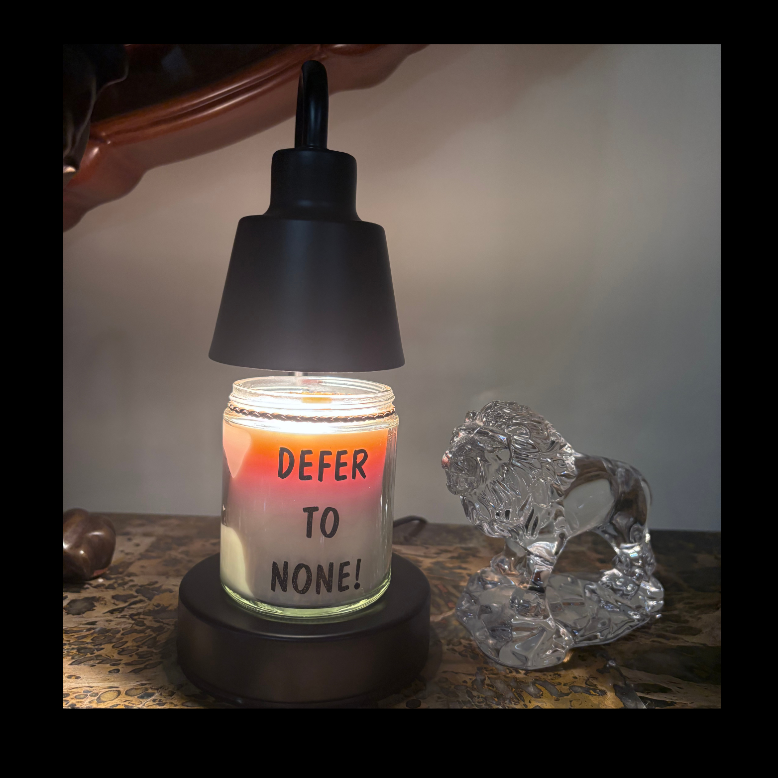 Candle with 'Defer to None!' text inside, lit by a black lamp, next to a crystal lion figurine.
