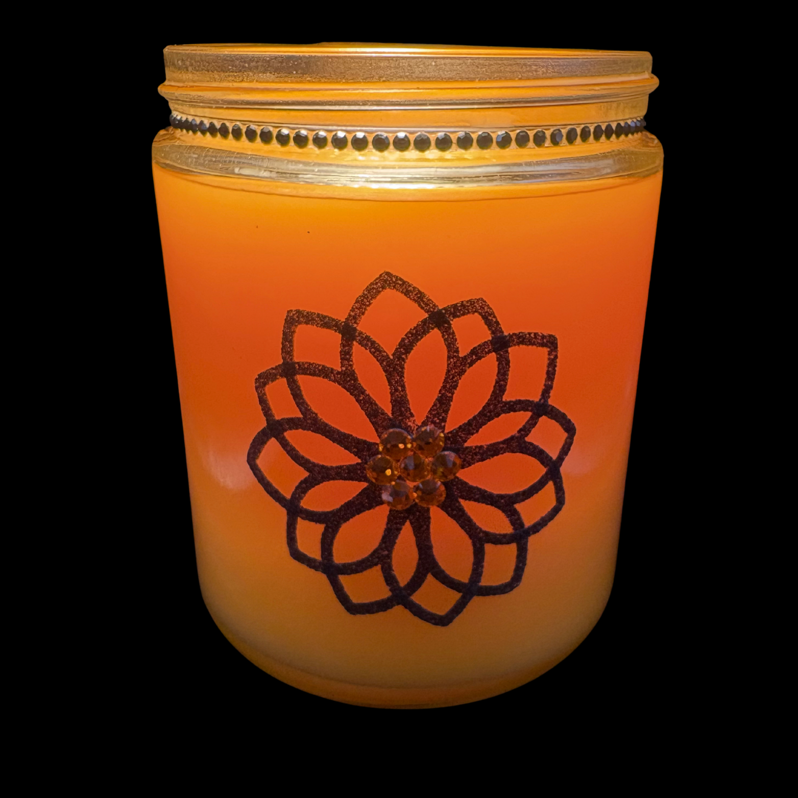 Orange candle in a glass jar with a decorative black flower design on a black background